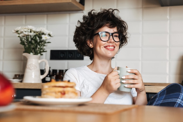 Mulher sorridente e alegre tomando um saboroso café da manhã enquanto está sentada na cozinha de casa, bebendo café