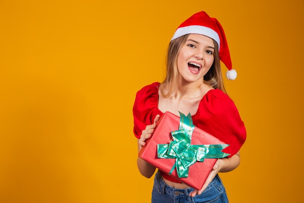 Mulher sorridente e animada com roupa vermelha de Papai Noel segurando o presente de Natal na orelha dela, isolado no fundo amarelo