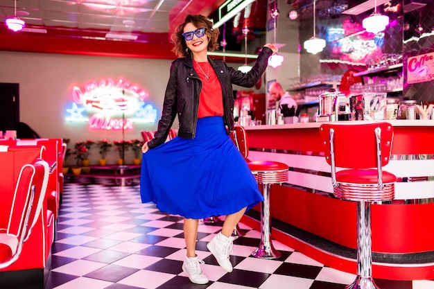 Mulher sorridente elegante com roupa colorida no café retrô dos anos 50 dançando vestindo jaqueta azul saia e óculos de sol de camisa vermelha se divertindo em clima alegre