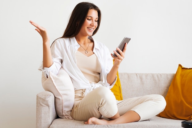 Mulher sorridente em camisa branca se senta no sofá branco, falando no smartphone e mostrando um gesto emocional com a mão.