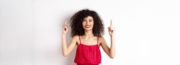 Mulher sorridente em elegante vestido vermelho e maquiagem apontando os dedos para cima e mostrando oferta promocional no valenti
