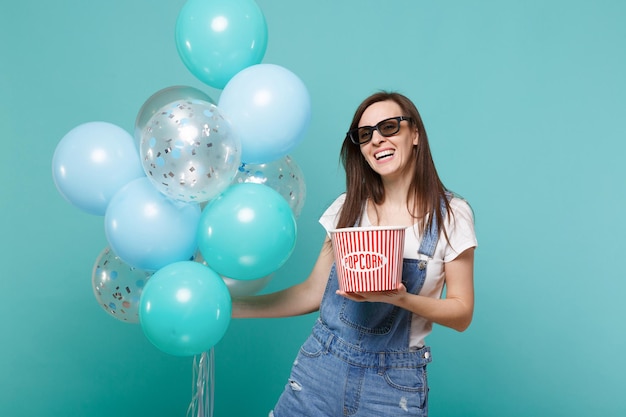 Mulher sorridente em óculos 3d assistindo filme filme segura balde de pipoca comemorando com balões de ar coloridos isolados em fundo azul turquesa. festa de aniversário, emoções no conceito de cinema.