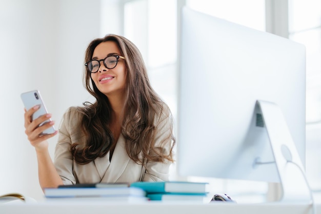 Mulher sorridente em roupas corporativas assiste a vídeos engraçados via smartphone senta-se na área de trabalho no escritório usa computador para trabalhar gosta de processo de trabalho navega informações para projeto usa óculos