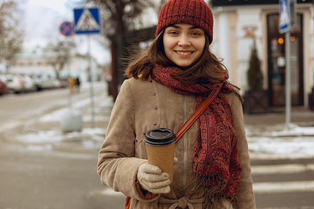 mulher sorridente em roupas quentes com xícara de papel em branco de café para viagem desfrutando de passeio na cidade
