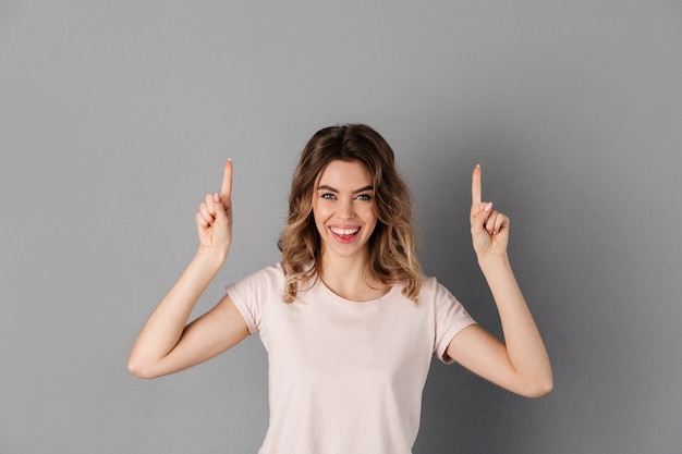 Mulher sorridente em t-shirt apontando para cima sobre cinza