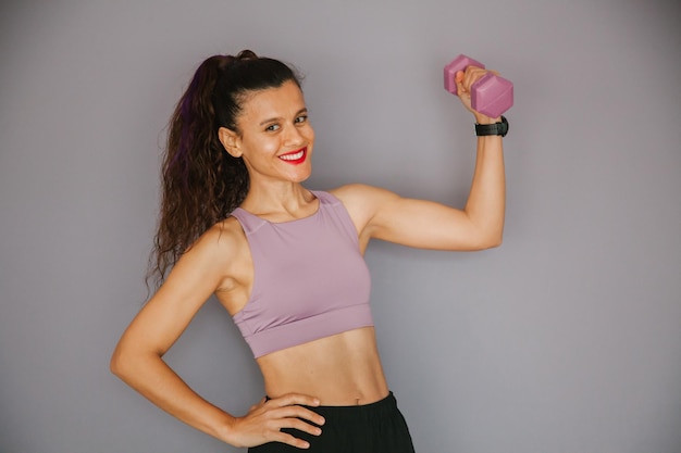 Mulher sorridente em um croptop roxo com um haltere na mão e um fundo roxo praticando esportes