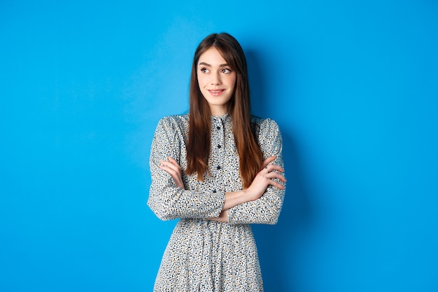 Mulher sorridente em vestido romântico cruza os braços no peito e olhando para a esquerda no logotipo, em pé contra o azul.