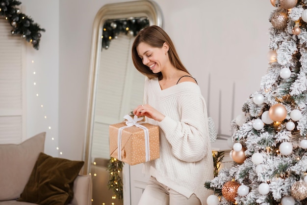 Mulher sorridente está abrindo a caixa de presente ao lado da árvore de Natal, dentro de casa.