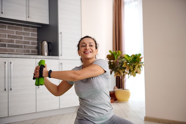 Mulher sorridente, esticando o corpo com halteres após o treino de ginástica em casa.