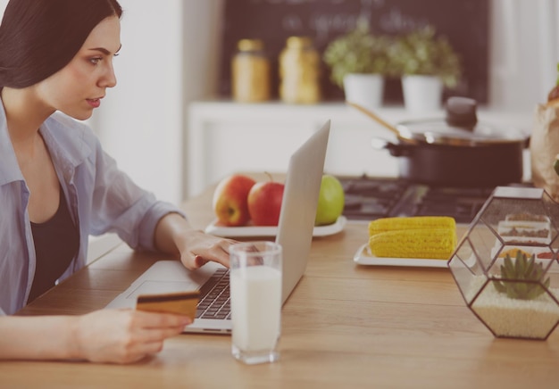 Mulher sorridente fazendo compras online usando computador e cartão de crédito na cozinha