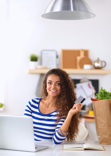 Mulher sorridente fazendo compras online usando computador e cartão de crédito na cozinha