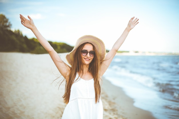 Mulher sorridente feliz em êxtase de felicidade livre na praia do oceano, de pé com um chapéu, óculos escuros e mãos levantadas. Retrato de um modelo feminino multicultural em vestido branco de verão curtindo a natureza durante o trav