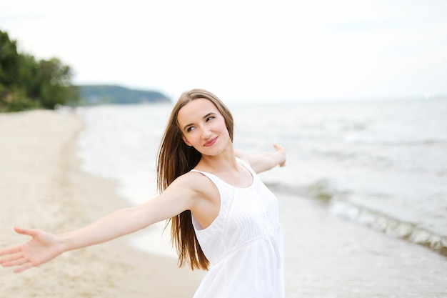 Mulher sorridente feliz em êxtase de felicidade livre na praia do oceano em pé com as mãos abertas. Retrato de um modelo feminino multicultural em vestido branco de verão curtindo a natureza durante as férias de viagem