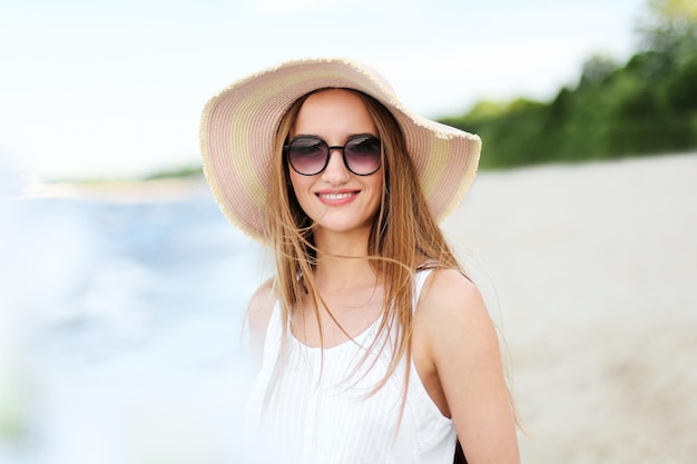 Mulher sorridente feliz em êxtase de felicidade livre na praia do oceano em pé com chapéu, óculos escuros e flores brancas. Retrato de um modelo feminino multicultural em vestido branco de verão curtindo a natureza durante a viagem