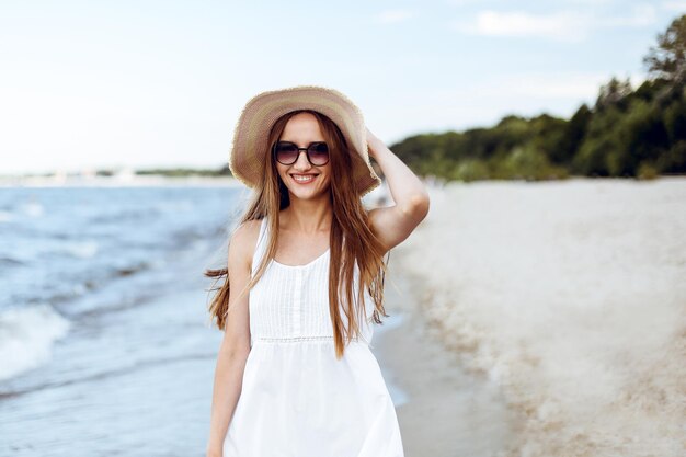 Mulher sorridente feliz em êxtase de felicidade livre na praia do oceano em pé e posando com chapéu e óculos de sol. Retrato de uma modelo feminina em vestido branco de verão curtindo a natureza durante as férias de viagem