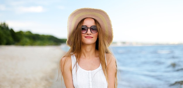 Mulher sorridente feliz em êxtase de felicidade livre na praia do oceano em pé e posando com chapéu e óculos de sol. Retrato de uma modelo feminina em vestido branco de verão curtindo a natureza durante as férias de viagem