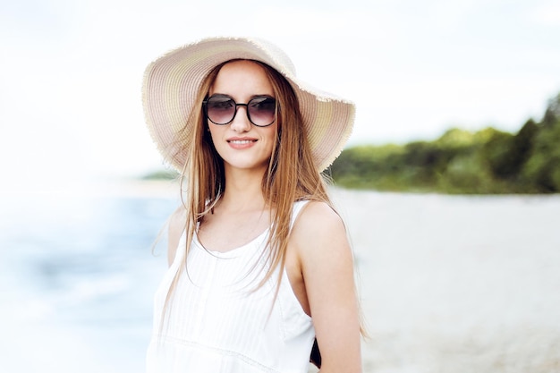 Mulher sorridente feliz em felicidade livre na praia do oceano de pé com chapéu, óculos de sol e flores brancas. Retrato de uma modelo feminina multicultural em vestido de verão branco desfrutando da natureza durante o trave