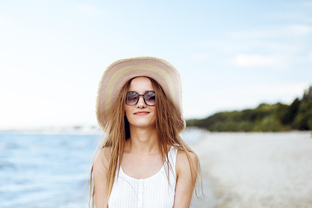Mulher sorridente feliz em felicidade livre na praia do oceano de pé e posando com chapéu e óculos de sol. Retrato de uma modelo feminina em vestido branco de verão desfrutando da natureza durante férias de viagem