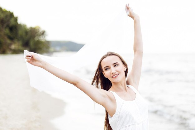 Mulher sorridente feliz em felicidade livre na praia do oceano pegando nuvens. Retrato de uma modelo feminina multicultural em vestido de verão branco desfrutando da natureza durante férias de viagem ao ar livre