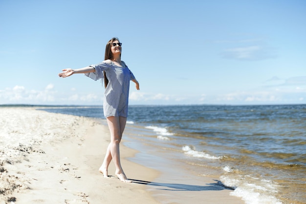 Mulher sorridente feliz em felicidade livre na praia oceânica em pé com as mãos abertas. Retrato de uma modelo feminina morena em vestido de verão apreciando a natureza durante as férias de viagem ao ar livre, plano geral