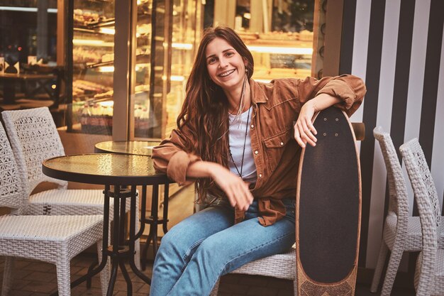 Mulher sorridente feliz está sentado no café do lado de fora enquanto segura seu longboard.