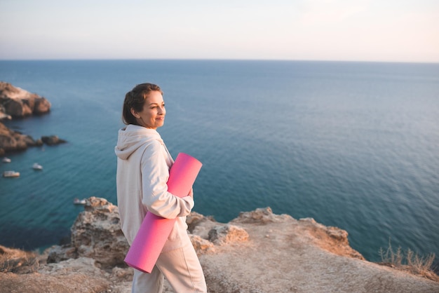 Mulher sorridente feliz segurando tapete de ioga rosa usar capuz esportivo e calças em cima da rocha sobre o fundo do lado do mar menina muito adulta pratica fitness ao ar livre fora estilo de vida saudável