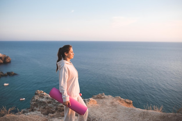 Mulher sorridente feliz segurando tapete de ioga rosa usar capuz esportivo e calças em cima da rocha sobre o fundo do lado do mar Menina muito adulta pratica fitness ao ar livre fora Estilo de vida saudável