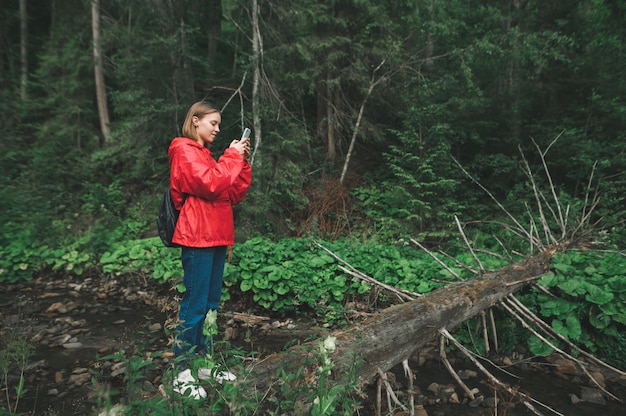 Foto mulher sorridente fica em pé sobre o córrego da montanha na floresta e usa smartphone