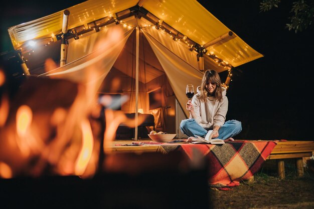 Mulher sorridente freelancer bebendo vinho e lendo livro sentado em aconchegante barraca de glamping na noite de outono Barraca de acampamento de luxo para férias ao ar livre e férias Conceito de estilo de vida