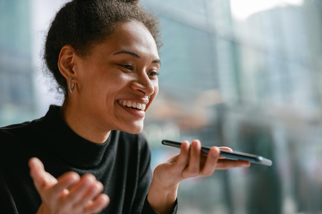 Mulher sorridente gravando mensagem de áudio no celular enquanto está sentada em um café aconchegante perto da janela