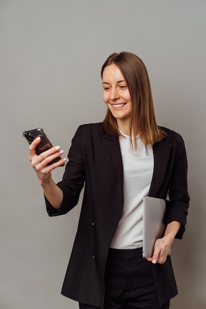 Mulher sorridente inteligente vestindo jaqueta está segurando um laptop e seu celular