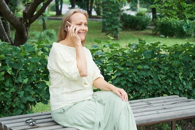 Mulher sorridente madura falando no celular enquanto está sentado no banco do parque