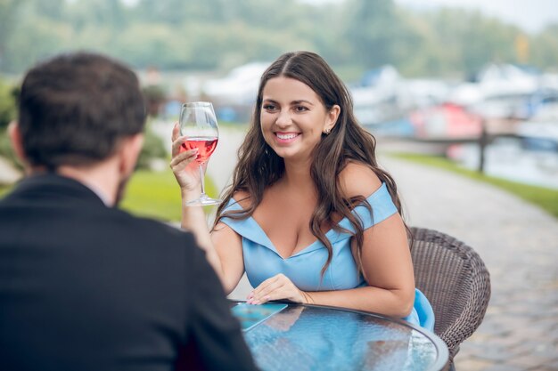 Mulher sorridente muito fofa de vestido azul e homem com taças de vinho em um café ao ar livre