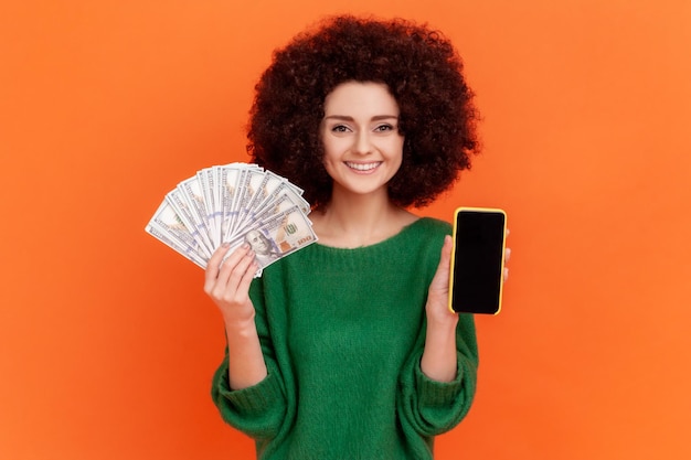 Mulher sorridente mulher com penteado afro vestindo camisola verde estilo casual segurando em mãos telefone inteligente com tela em branco e notas de dólares. tiro de estúdio interior isolado em fundo laranja.