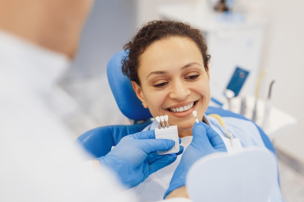 Mulher sorridente na cadeira odontológica escolhendo a cor dos dentes com dentista masculino em clínica privada