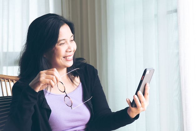 Mulher sorridente, negra, cabelo comprido, sentada, segurando um smartphone e se comunicando com a família