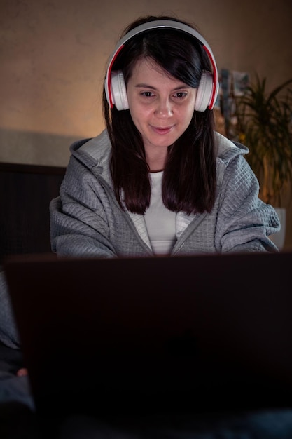 Mulher sorridente no fone de ouvido trabalhando no laptop sentado na cama