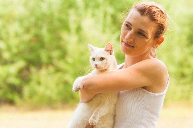 Mulher sorridente, olhando para a câmera segurando o gato fofo branco em seus braços em pé no parque de verão