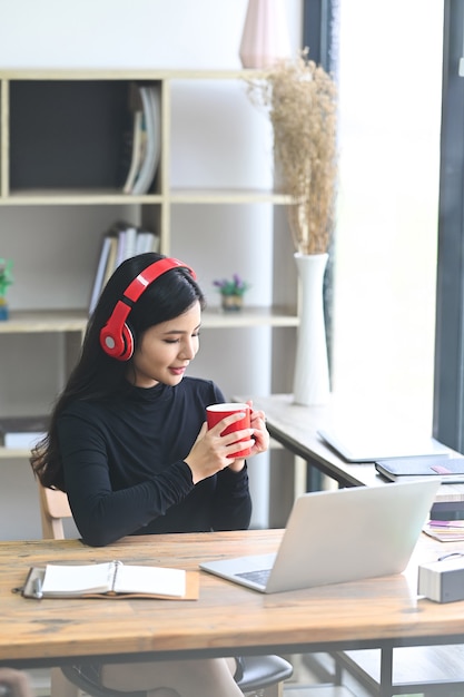 Mulher sorridente, ouvindo música no fone de ouvido e bebendo café no escritório.