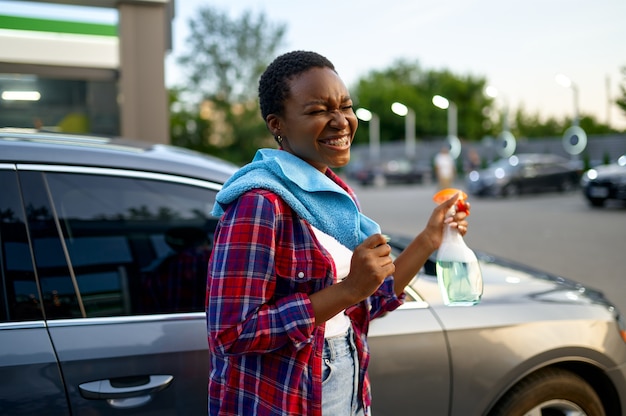 Mulher sorridente posa com spray limpador de janela e um pano, estação de lavagem de carros de mão. Indústria ou negócio de lavagem de carros. Mulher limpa seu veículo de sujeira ao ar livre