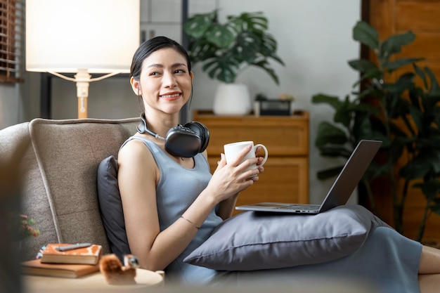 Mulher sorridente relaxando em casa usando laptop ouvindo música e bebendo café quente