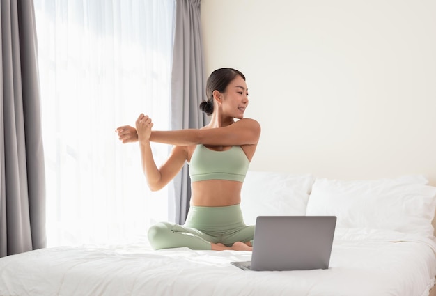 Mulher sorridente se alongando e se preparando para fazer ioga na cama durante o treinamento online pela manhã em casa