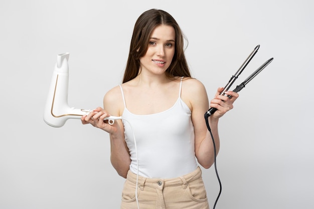 Mulher sorridente segurando alisador de cabelo e secador de cabelo isolado no fundo branco Beleza