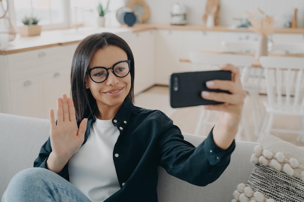 Mulher sorridente segurando o telefone acenando com a mão conversando por videochamada sentada no sofá em casa