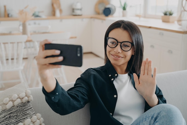 Mulher sorridente segurando o telefone acenando com a mão conversando por videochamada sentada no sofá em casa