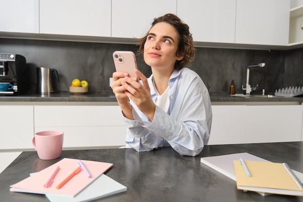 Mulher sorridente segurando smartphone em sua cozinha usando telefone celular e parecendo feliz