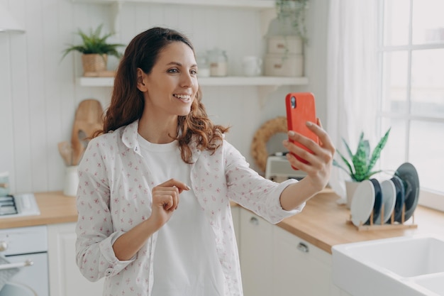 Mulher sorridente segurando telefone fazendo videochamada desfrutando de conversa online na cozinha em casa