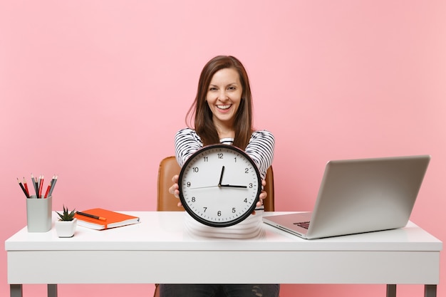 Mulher sorridente segurando um despertador redondo enquanto está sentado e trabalhando no projeto na mesa branca com um laptop pc contemporâneo