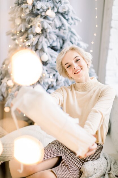 Mulher sorridente segurando uma caixa de presente sentada em uma sala decorada com árvore de natal