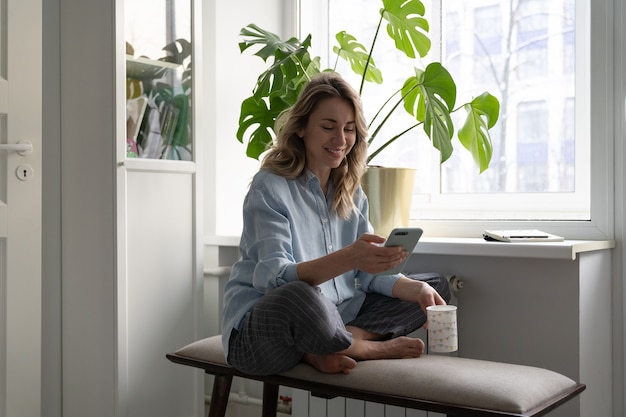 Mulher sorridente segurando uma xícara de café em casa, usando telefone celular inteligente, batendo papo nas redes sociais